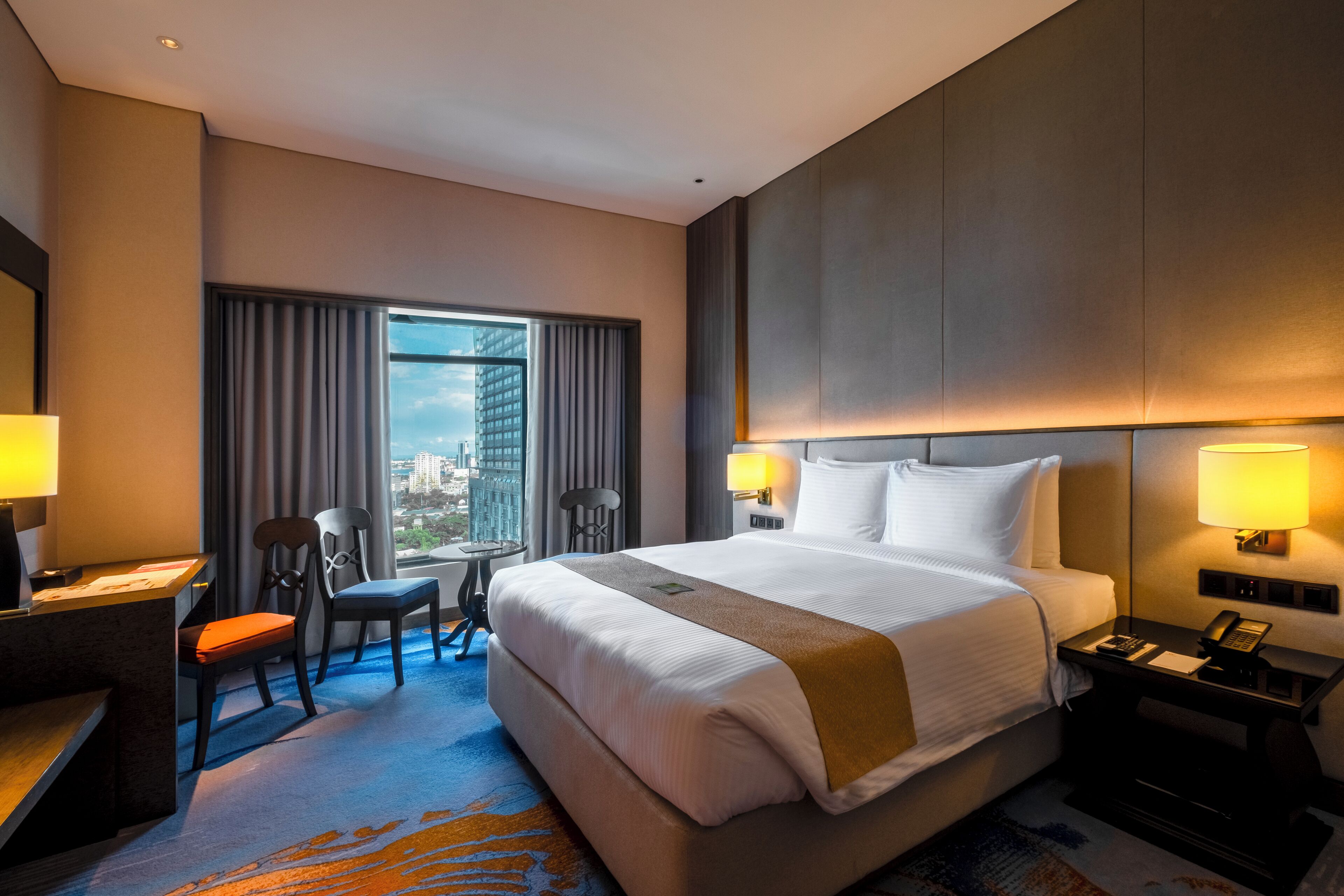 A hotel room with a large bed, two bedside lamps, a desk with a chair, and a view of the city through the window.