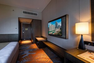 Habitación de hotel con cama, escritorio, lámpara, televisor mostrando un paisaje urbano y alfombra con diseño geométrico.