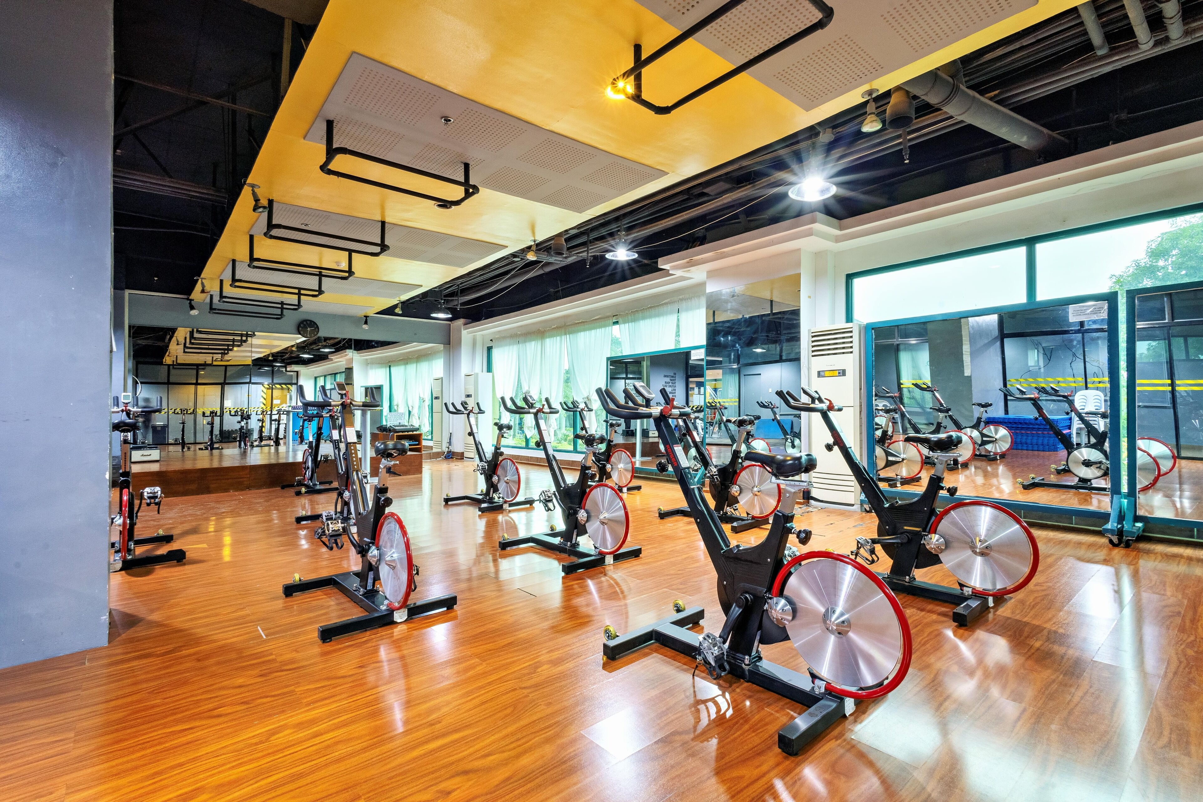 A gym with multiple stationary bikes arranged in rows, large windows, and a wooden floor.