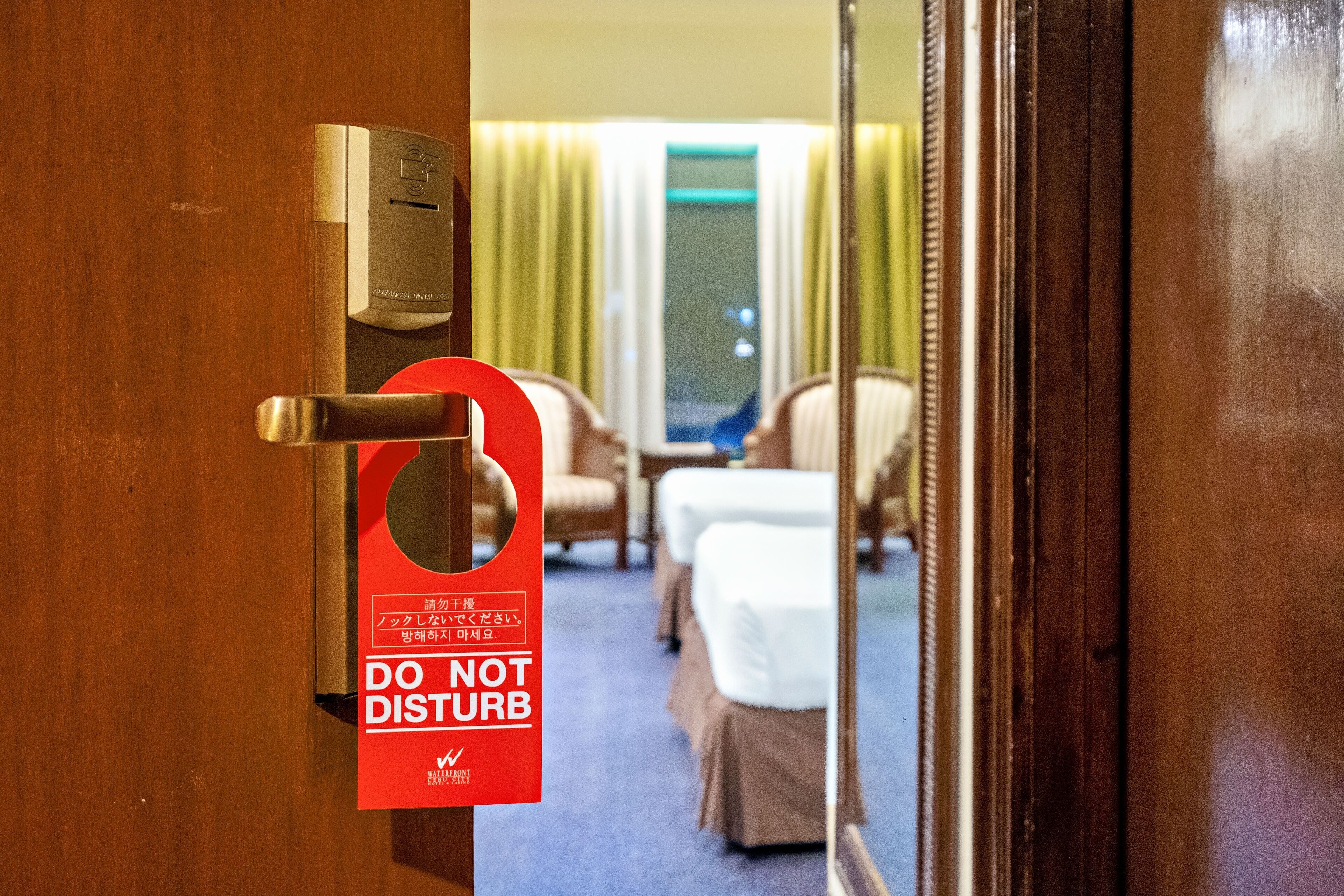 A hotel room door with a 'DO NOT DISTURB' sign, a keycard reader, and a view of the room's bed and chairs.