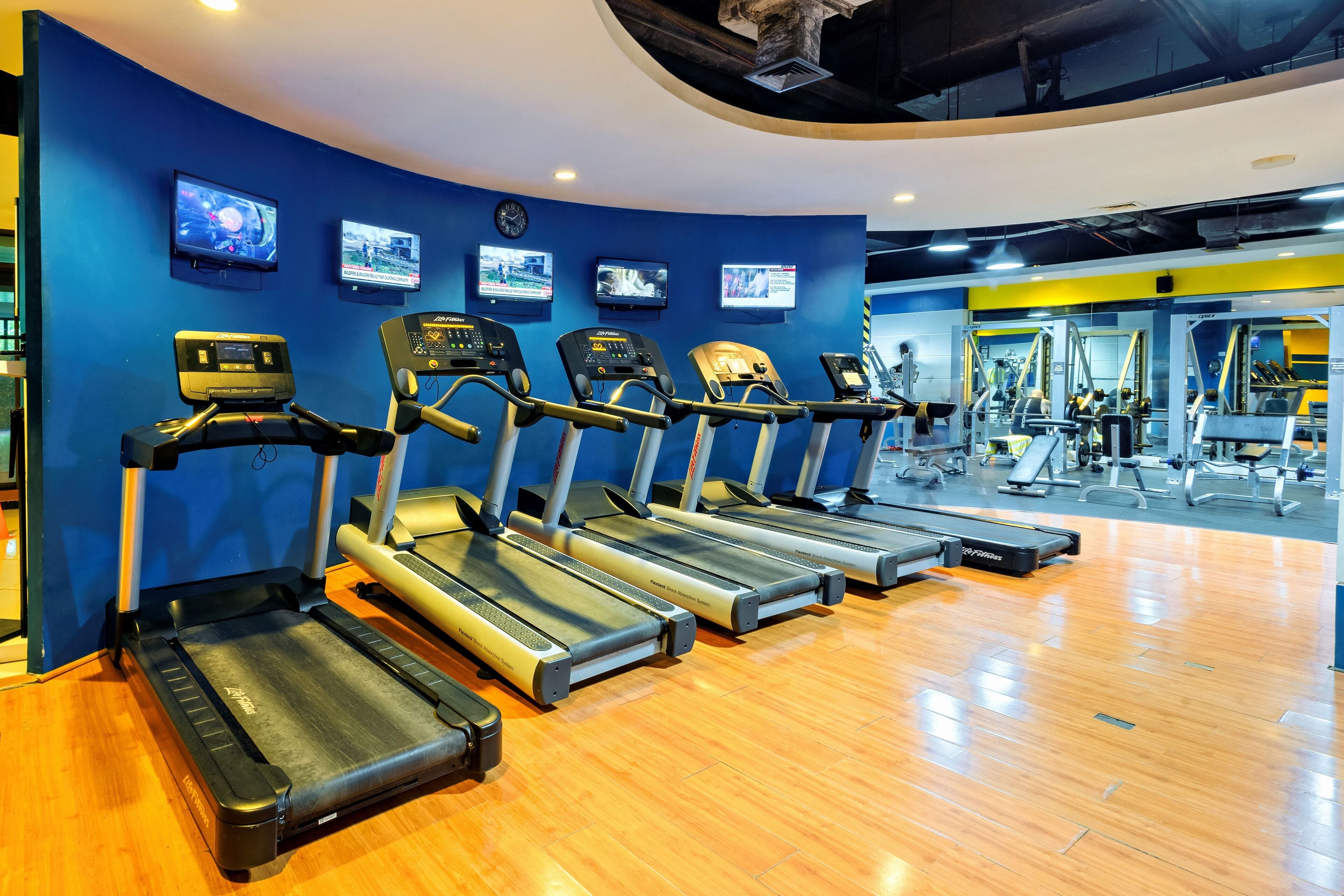 Un gimnasio con cintas de correr alineadas frente a una pared azul, un reloj y equipo de ejercicio al fondo.