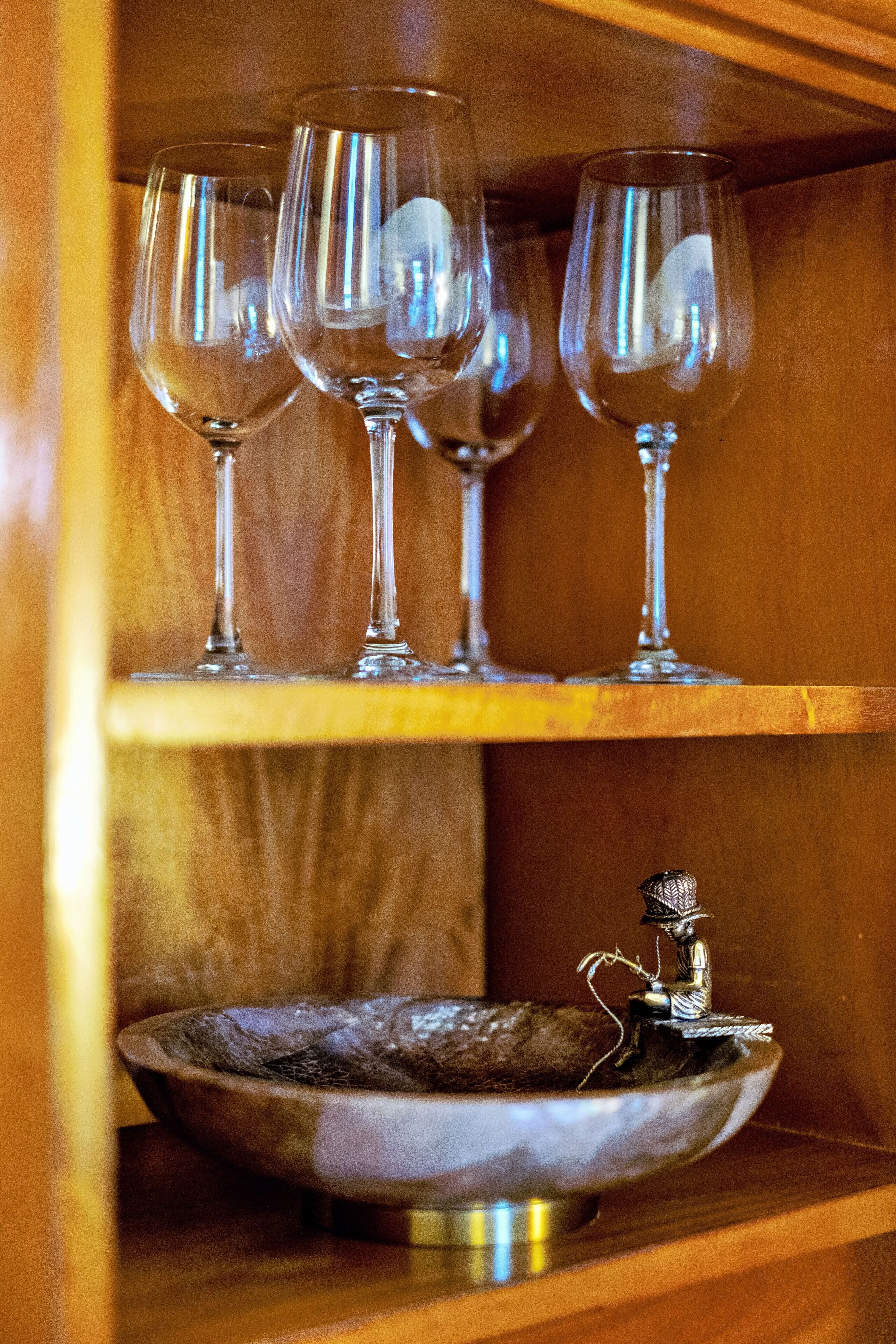 A wooden shelf with three wine glasses and a decorative metal bowl with a figure.