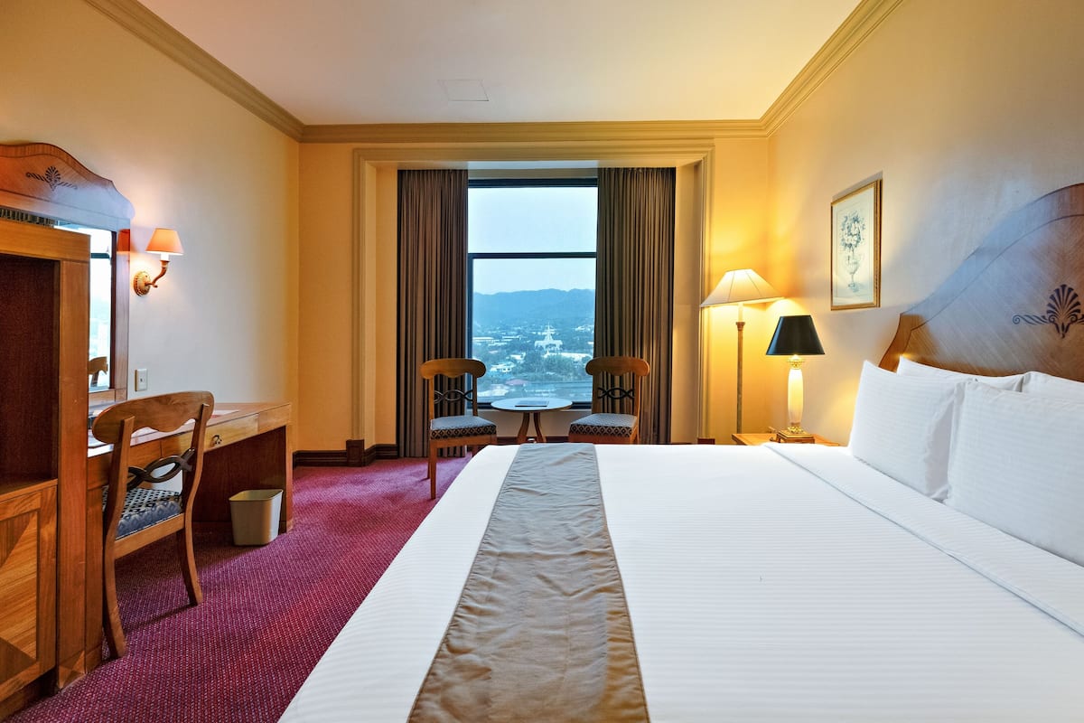 Une chambre d’hôtel avec un grand lit, un bureau, deux chaises, une petite table et une vue sur l’extérieur grâce à une grande fenêtre.