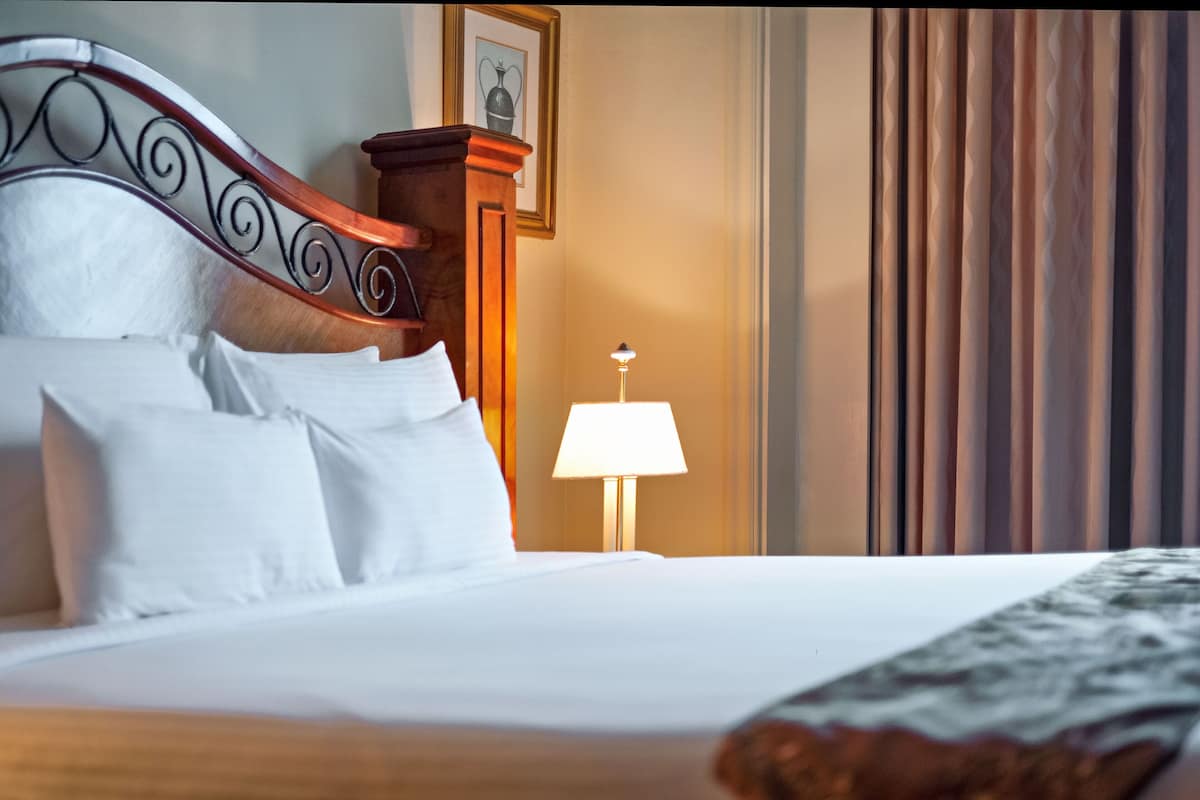 Un lit bien fait, avec du linge de lit blanc, une tête de lit en bois ornée d’un motif métallique décoratif, une lampe de chevet et un tableau encadré accroché au mur.