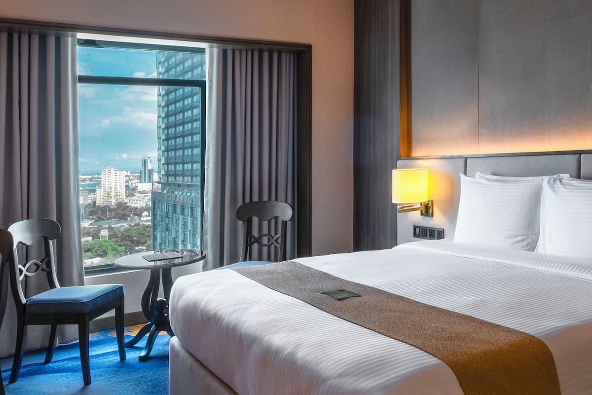 Une chambre d’hôtel avec un grand lit, une chaise, une petite table et une vue sur la ville par une grande fenêtre.