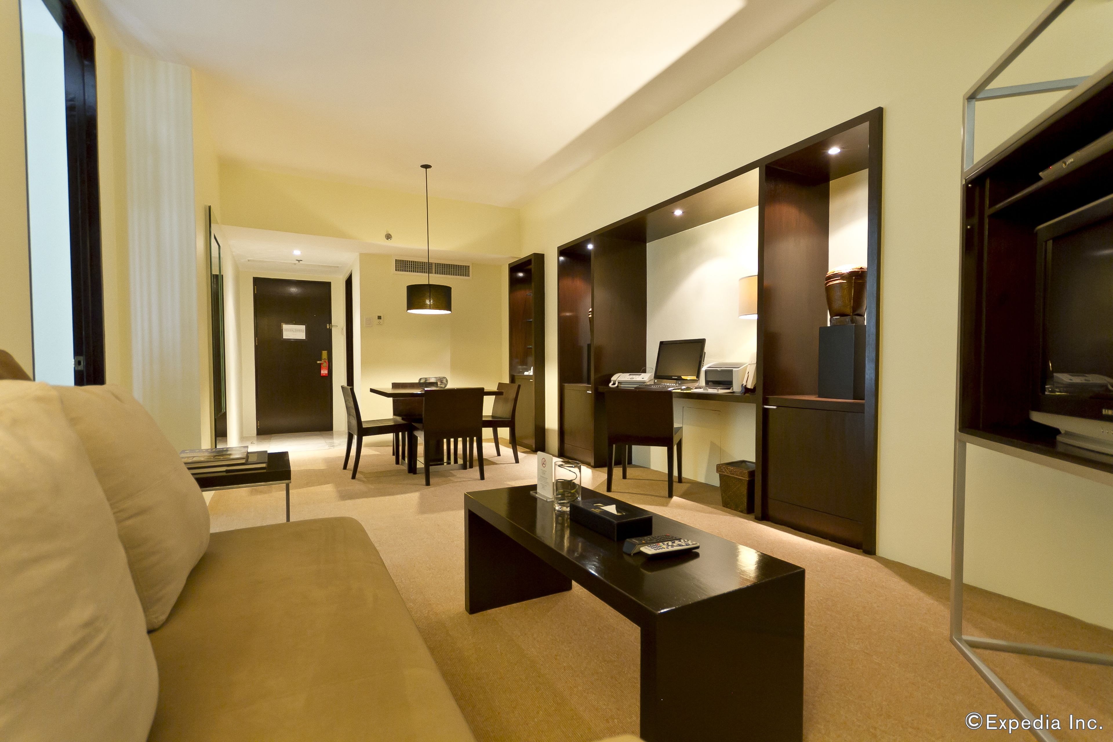 A modern hotel room with a sofa, a coffee table, a dining area with chairs, a kitchen area, and a TV.