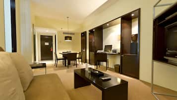 A modern hotel room with a sofa, a coffee table, a dining area with chairs, a kitchen area, and a TV.