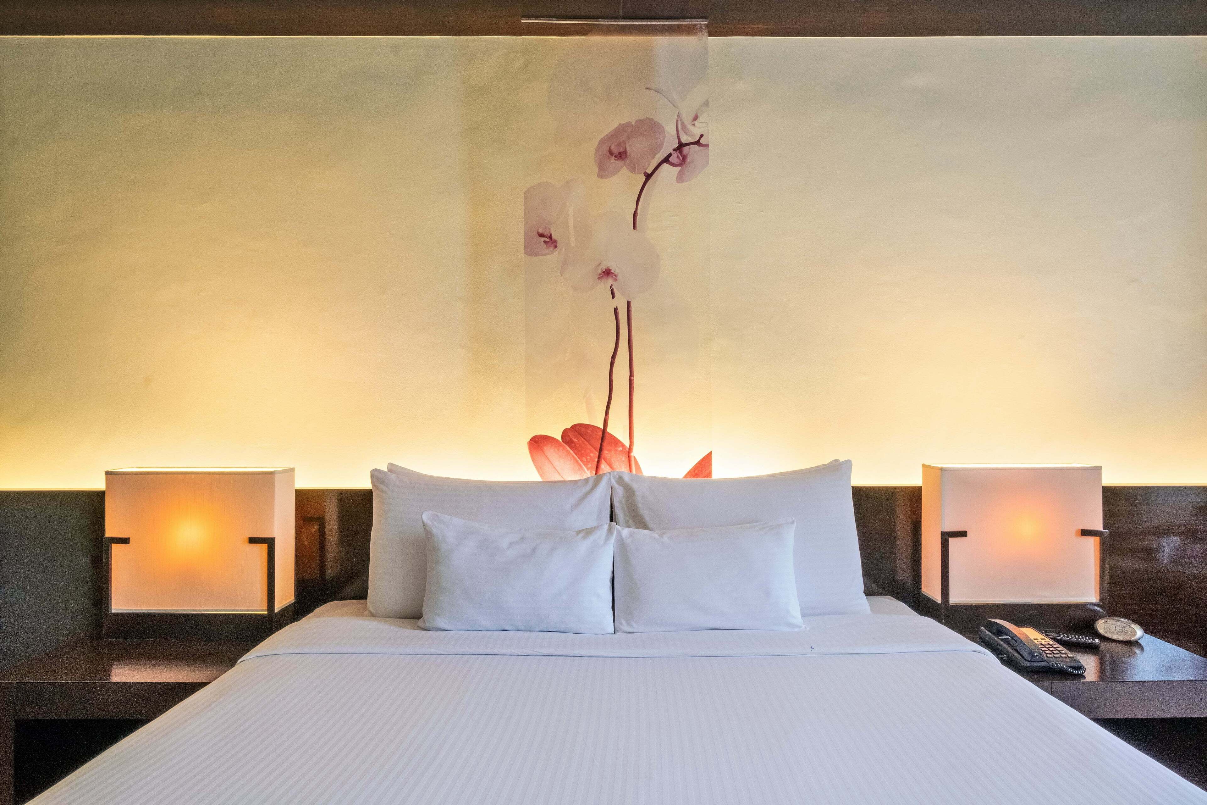 A hotel room with a large bed, two bedside tables, a nightstand, a wall-mounted telephone, and a decorative orchid wall hanging.