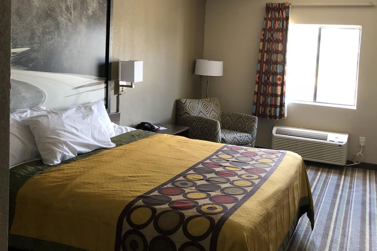 Une chambre d’hôtel comprenant un lit, un bureau, une chaise, une lampe, une fenêtre avec des rideaux et un appareil de climatisation.