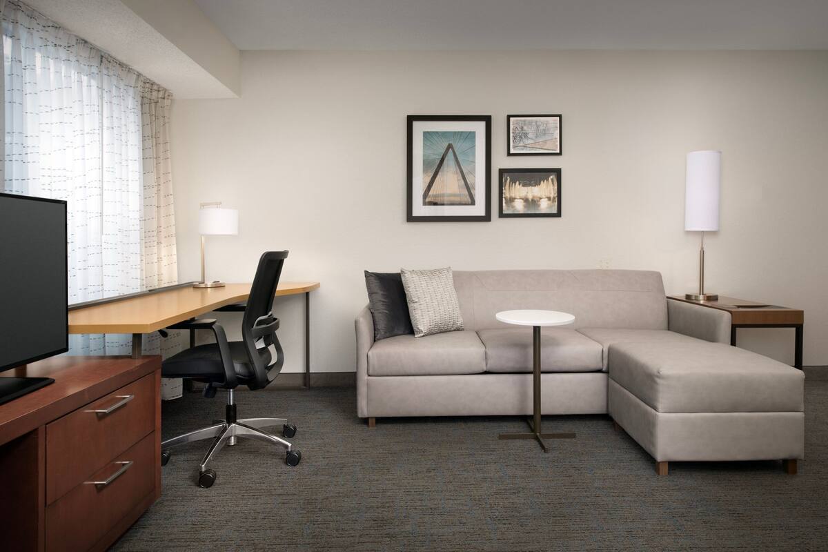 A modern hotel room with a desk, office chair, sofa, and a small round table.