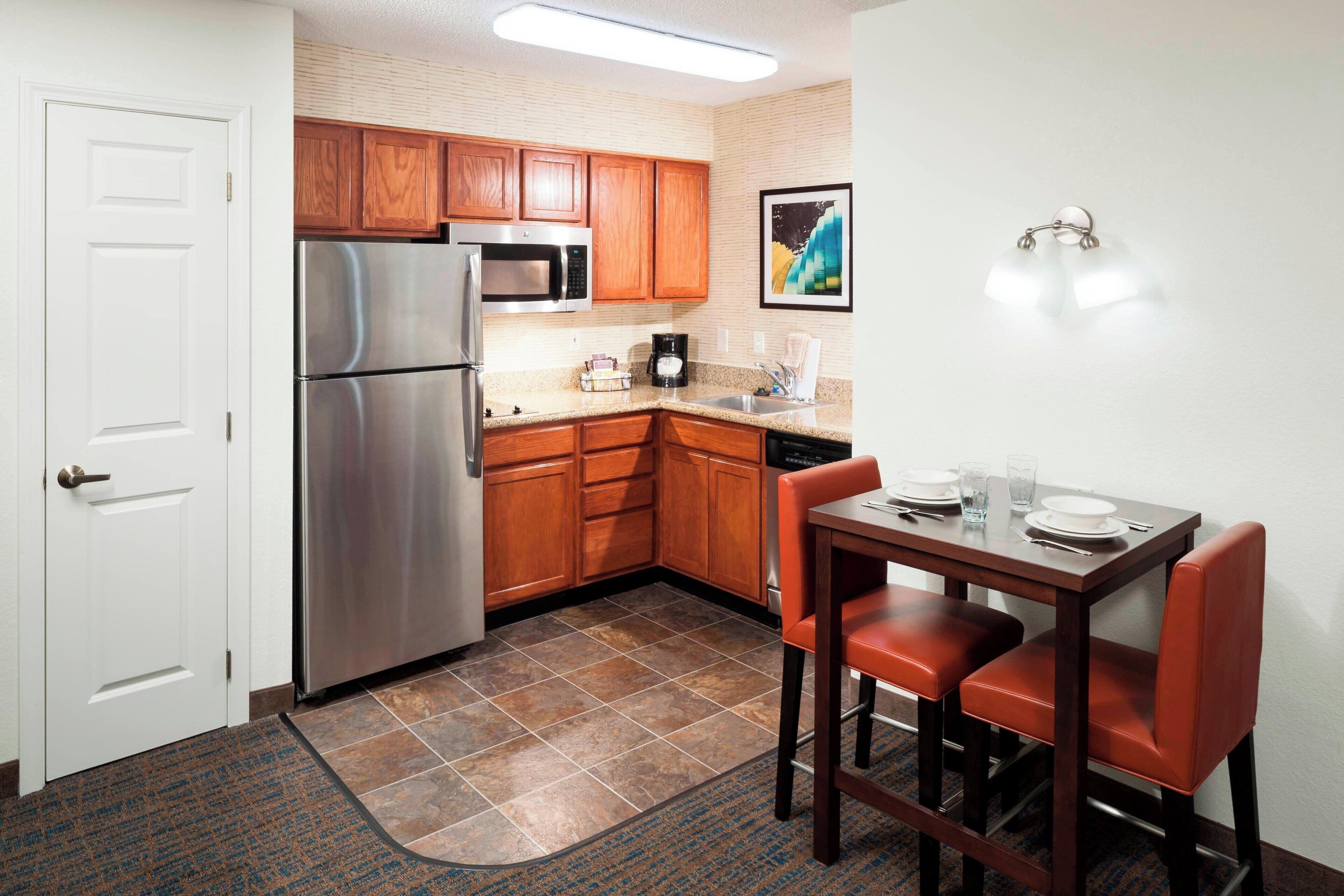 A compact kitchen with wooden cabinets, a stainless steel refrigerator, a microwave, and a dining table with two chairs.