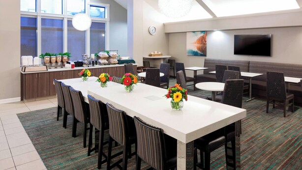 A modern dining area with a long table, chairs, and a kitchen counter with potted plants. - Residence Inn by Marriott Kansas City Overland Park (Overland Park)