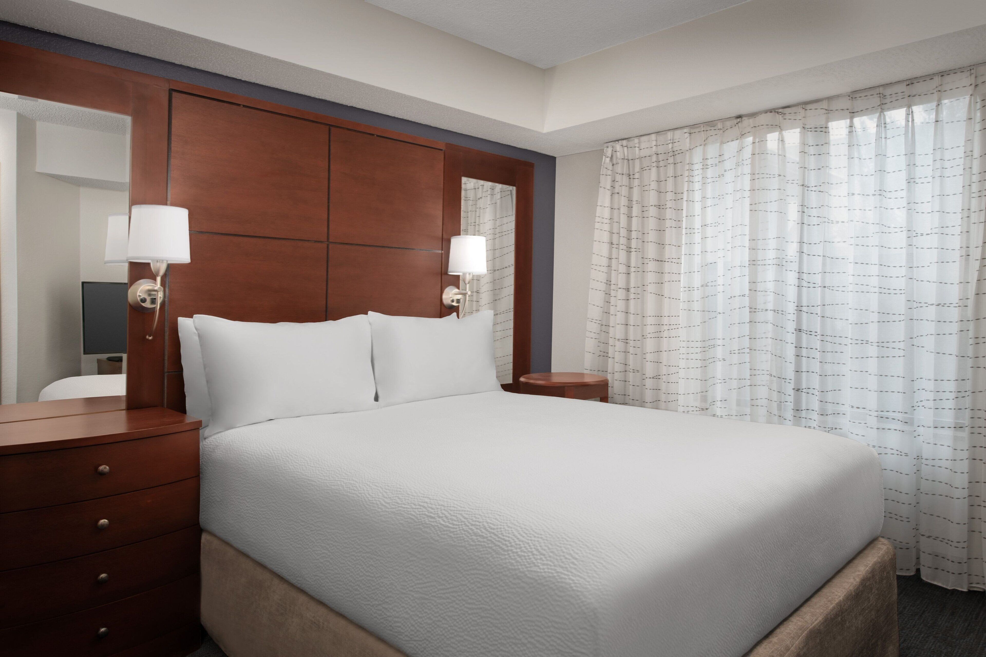 A neatly made bed with white linens, a wooden headboard, and two bedside lamps.