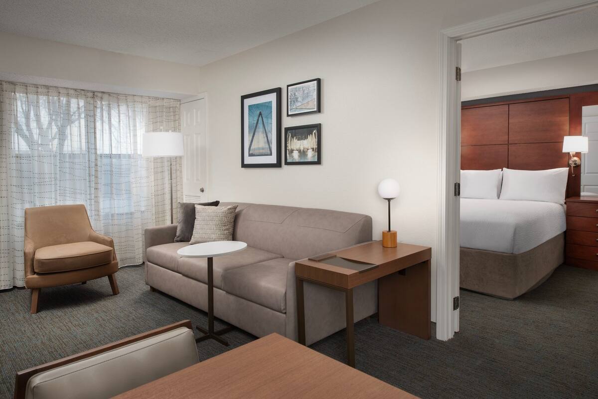 A hotel room with a sofa, armchair, small round table, and a bed with a wooden headboard. There are three framed pictures on the wall.
