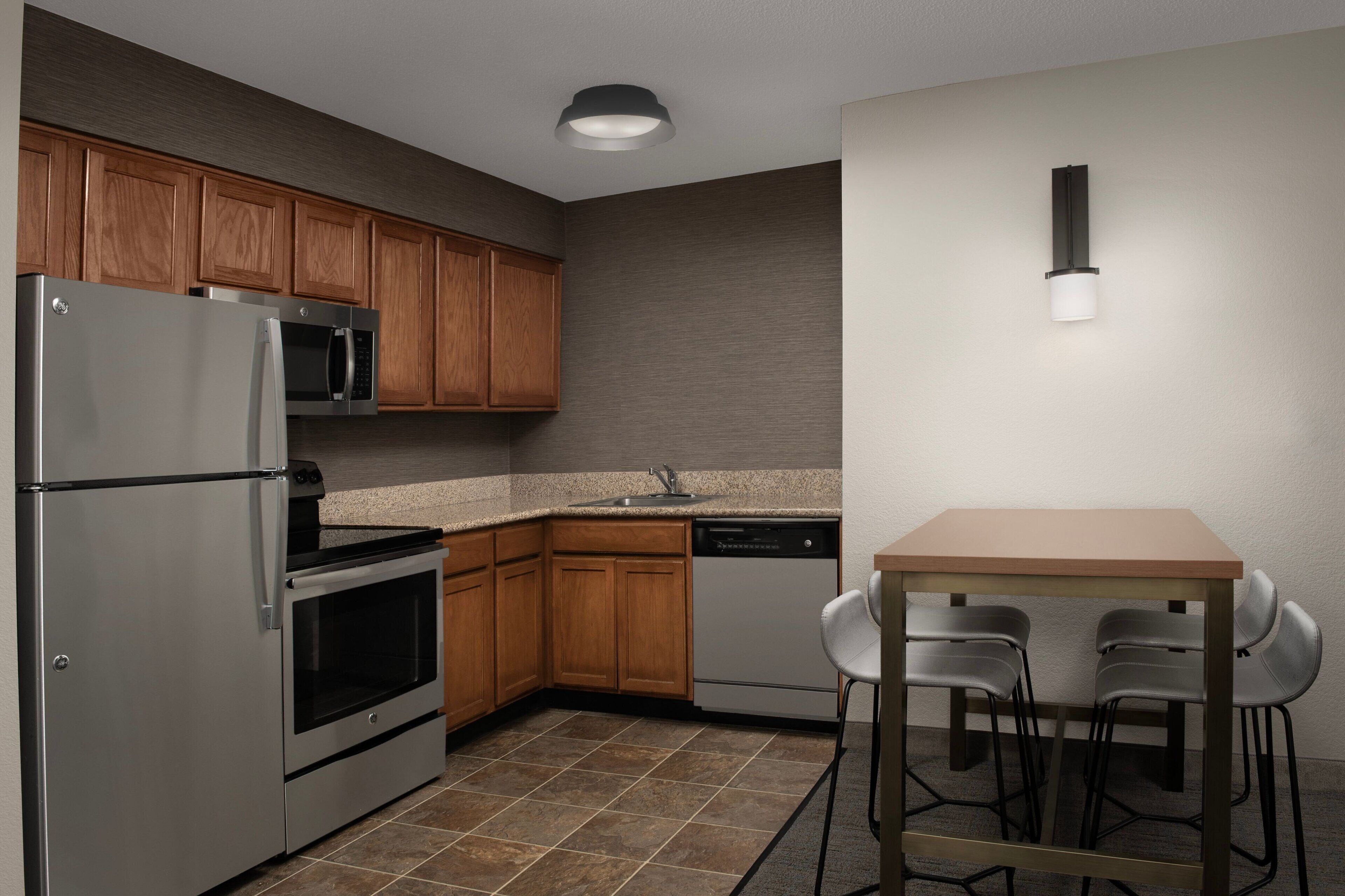 A modern kitchen with wooden cabinets, stainless steel appliances, a granite countertop, and a small dining area with a table and chairs.