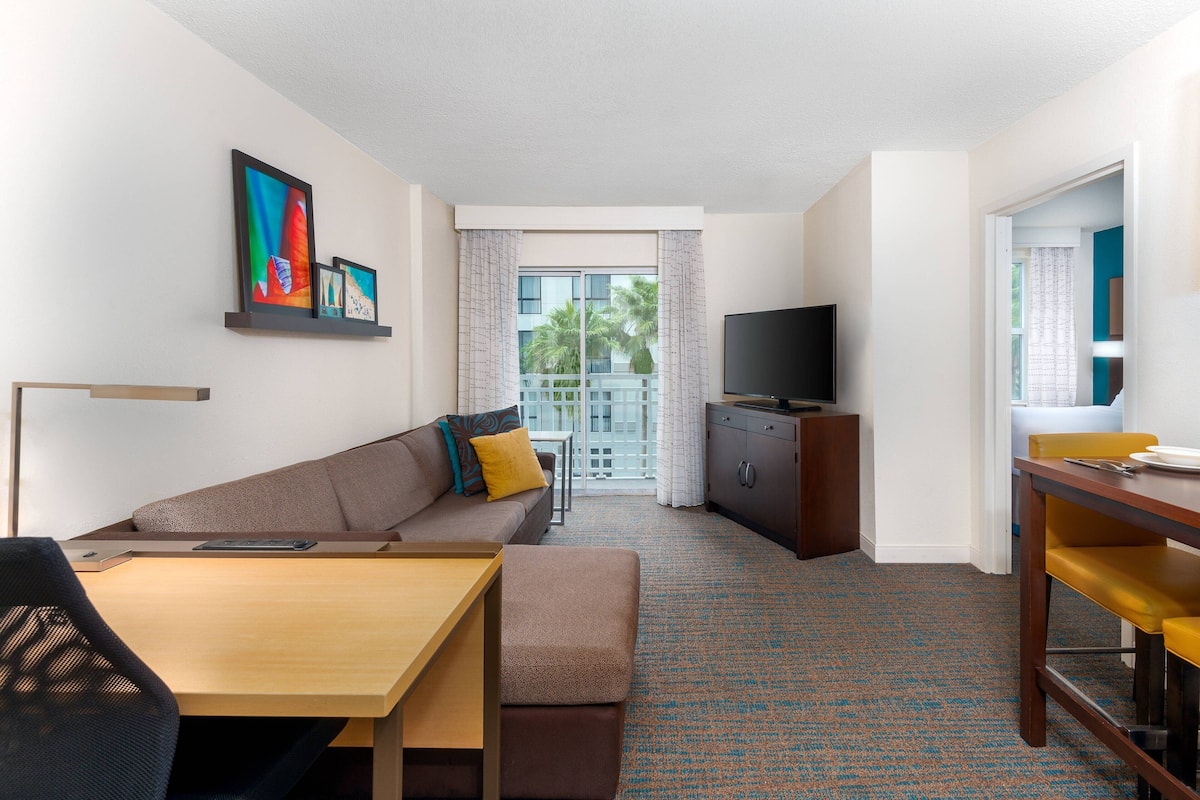 Habitación de hotel con sofá, mesa de comedor, televisor y balcón con vista a palmeras.