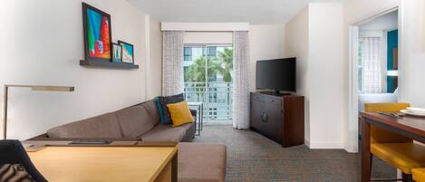 A hotel room with a sofa, a dining table, a TV, and a balcony with a view of palm trees.