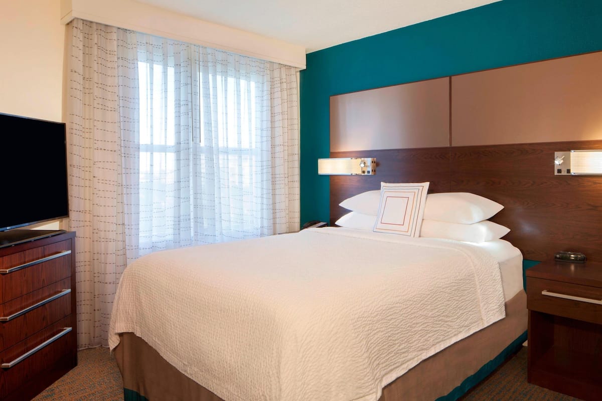 Una habitación de hotel con una cama grande, un televisor sobre un tocador de madera, una mesita de noche con una lámpara y una ventana con cortinas.