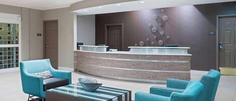 A modern reception area with a curved reception desk, teal armchairs, and a glass coffee table.