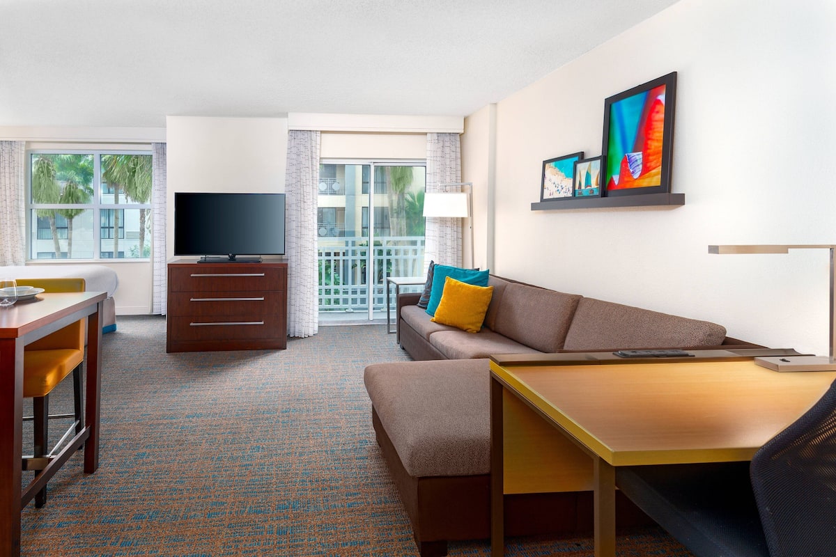 Habitación de hotel con sofá, televisión, escritorio y balcón con vista a palmeras.