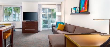 A hotel room with a sofa, a TV, a desk, and a balcony with a view of palm trees.