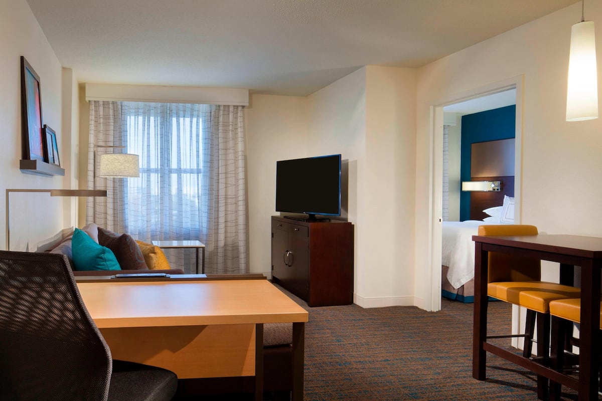 Habitación de hotel con cama, televisión sobre un mueble de madera, escritorio con silla y una pequeña zona bar.