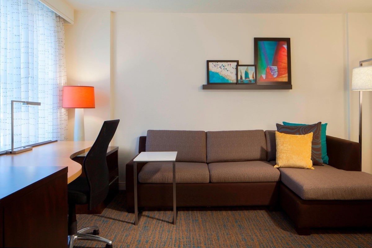 Una habitación de hotel moderna con un sofá marrón, una mesita pequeña, un escritorio y una lámpara. Hay pinturas abstractas en la pared.