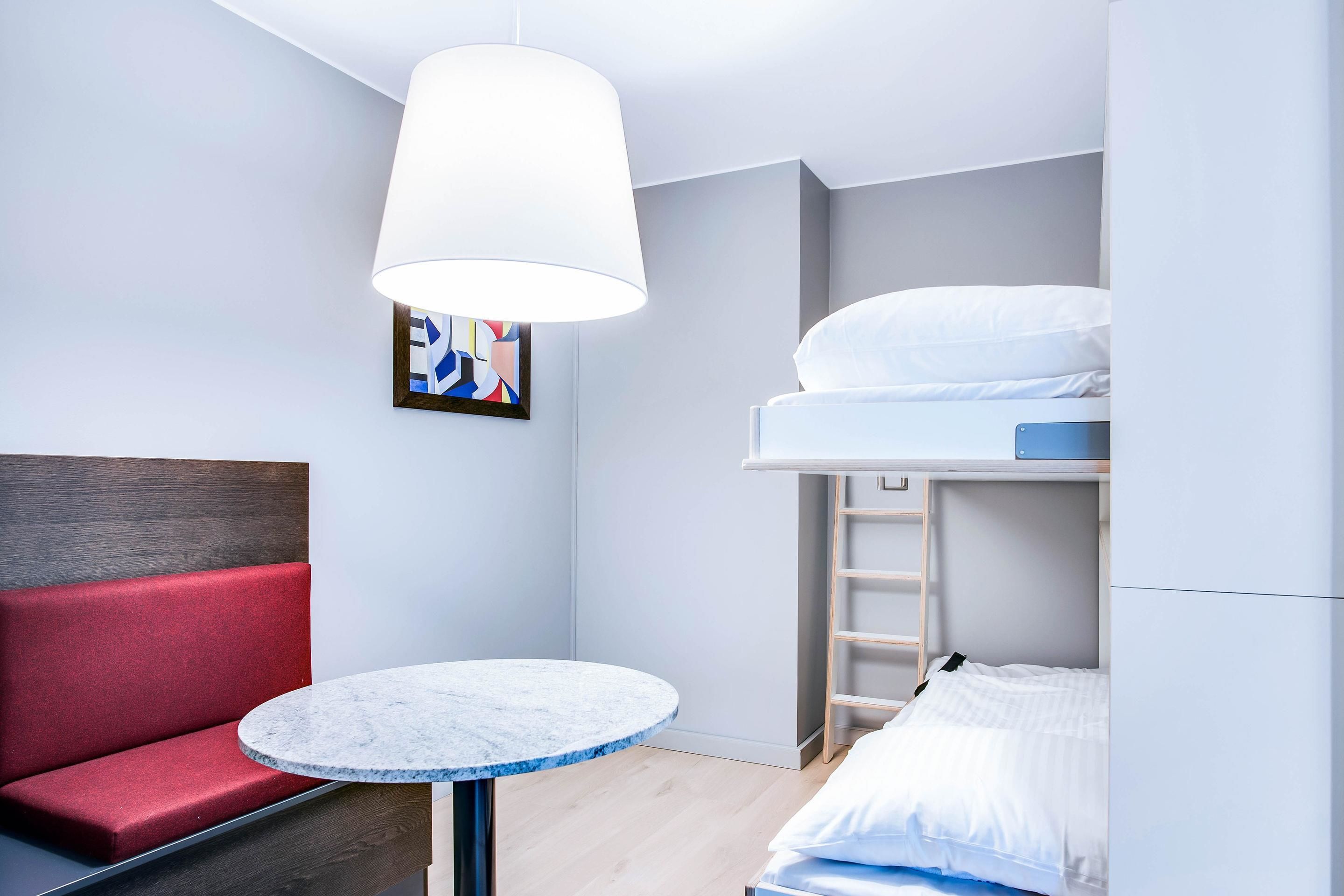 A compact room with a bunk bed, a round marble table, a red cushioned bench, and a white pendant light.