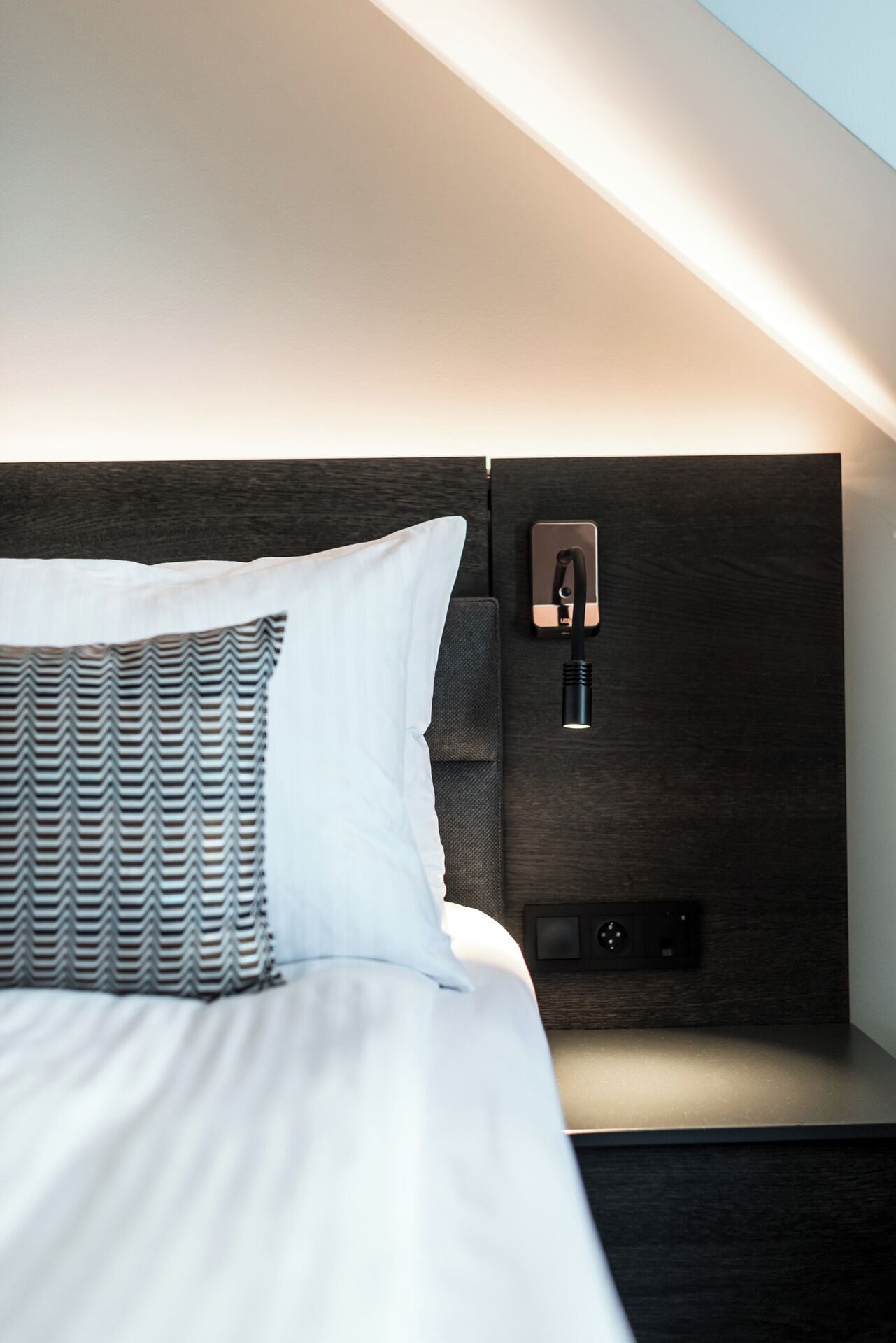 A modern hotel room with a neatly made bed, a nightstand with a lamp, and a wall-mounted charging station.