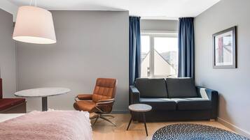 Ein modernes Hotelzimmer mit Bett, einer Couch, einem Sessel, einem kleinen Tisch und einem großen Fenster mit Vorhängen.