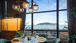A dining area with a table set for four, large windows with a view of the sea, and a hanging light fixture with multiple bulbs.