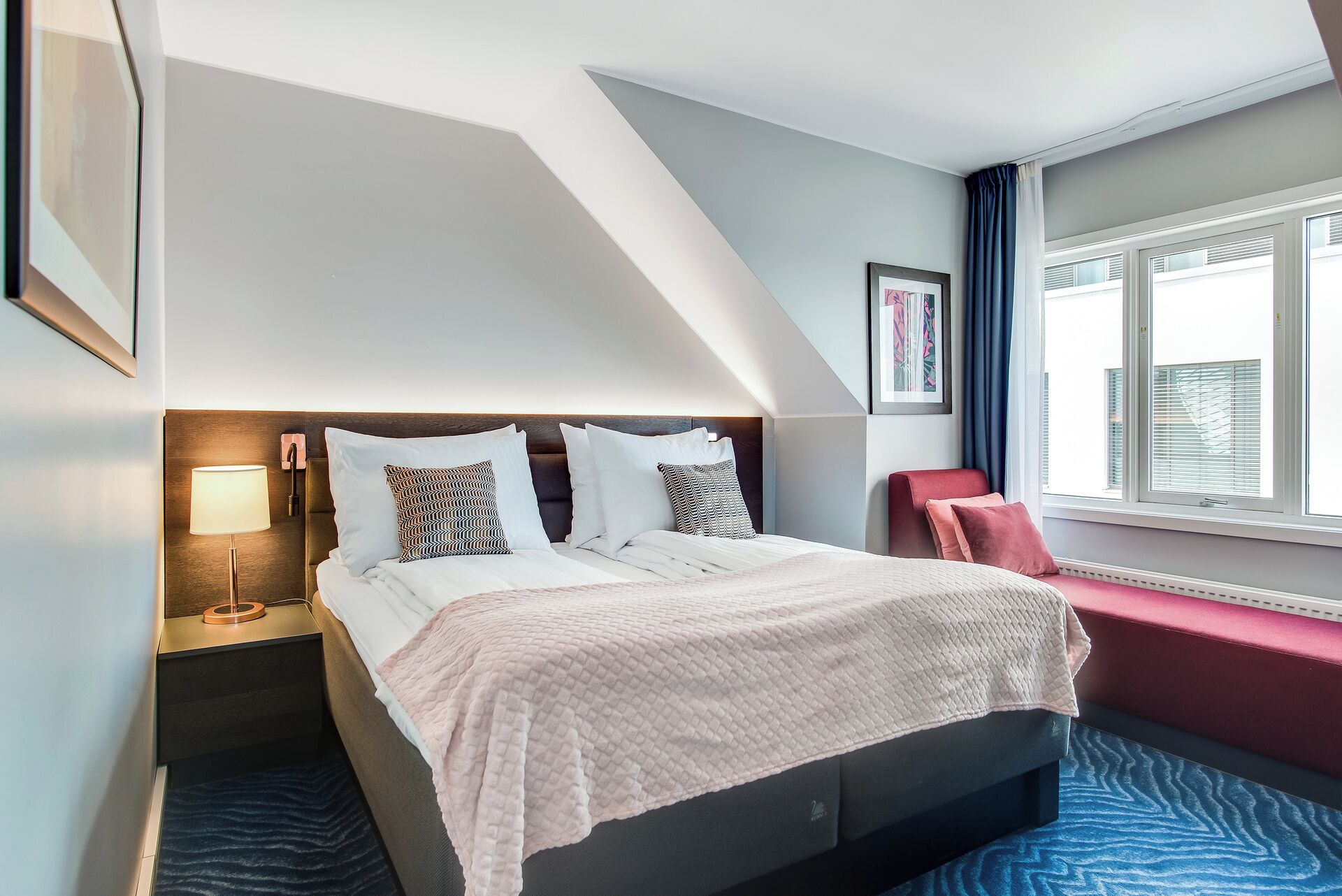 A hotel room with a bed, bedside table, lamp, and a window with blinds.