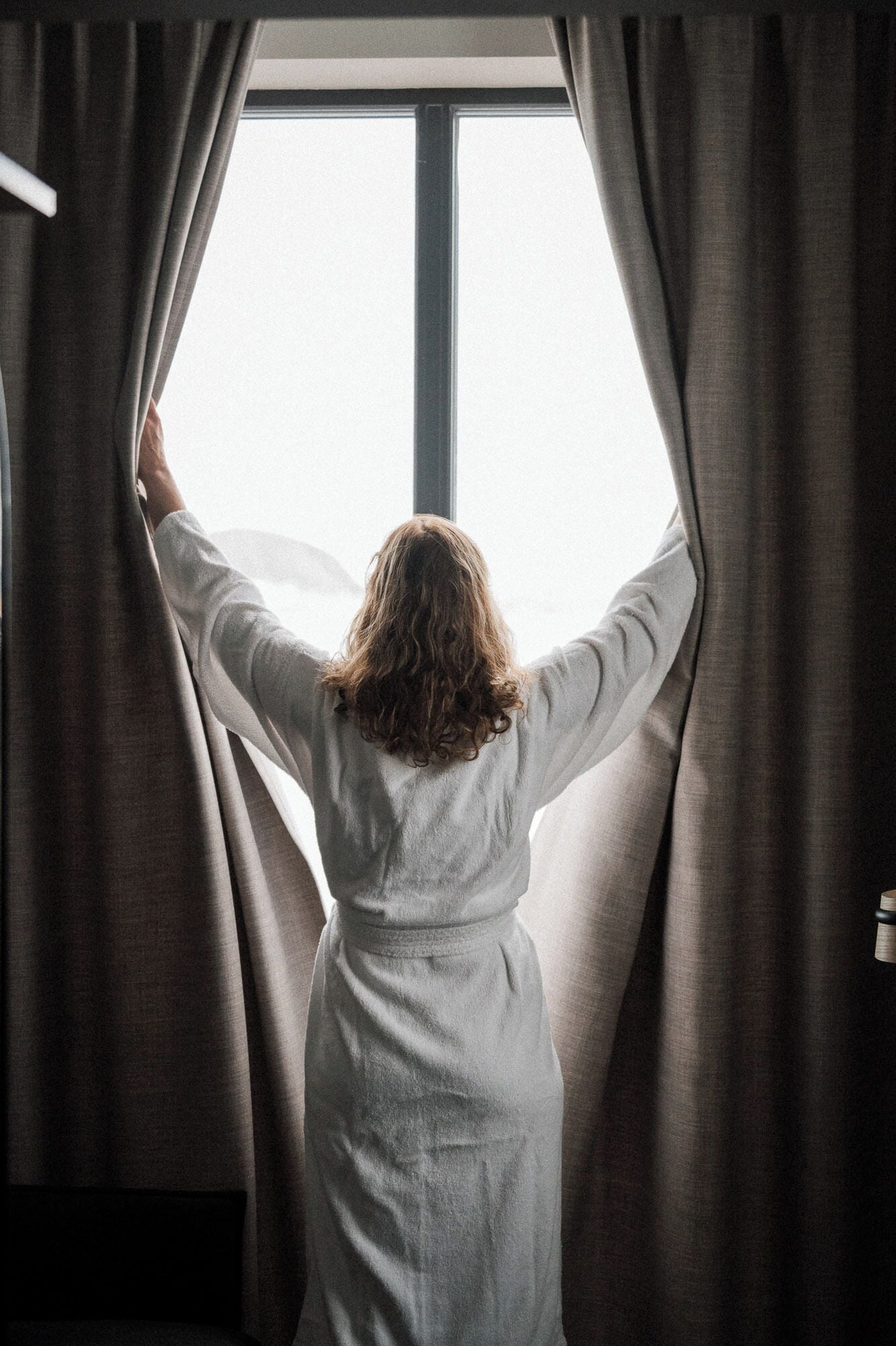 A person standing in a room with a large window, curtains partially drawn, and a visible door handle.