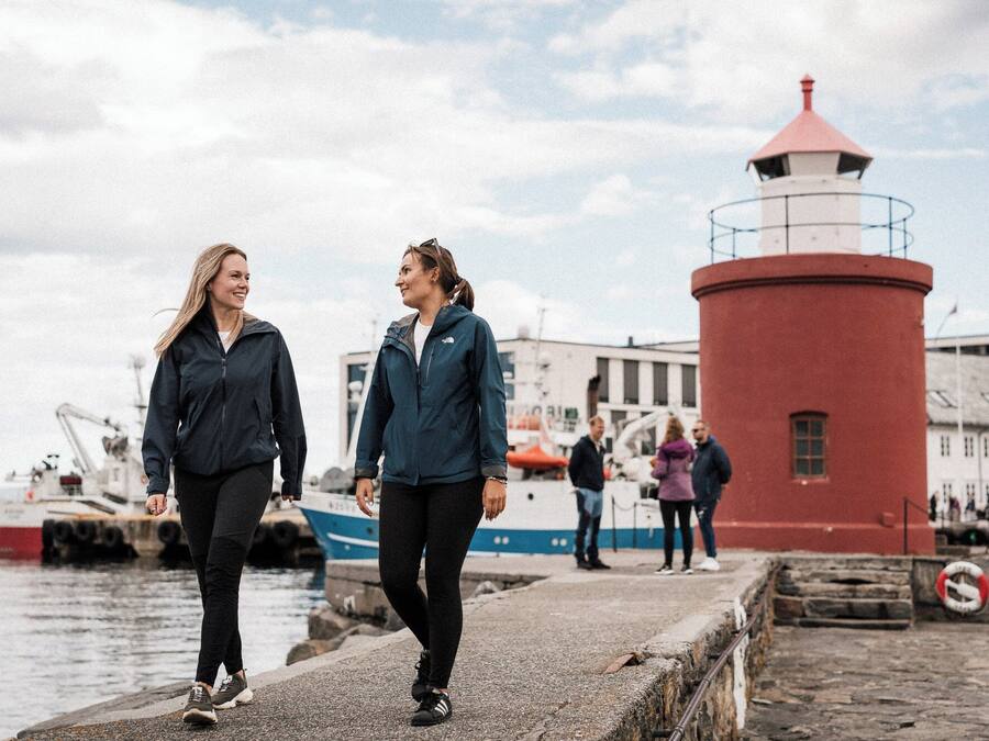 Two people walking on a pier with a red lighthouse in the background.