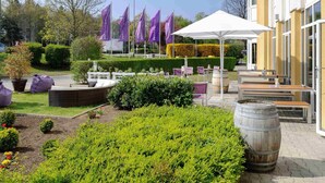 An outdoor seating area with tables and chairs, surrounded by greenery and a building with large windows. - Mercure Hotel Stuttgart Gerlingen (Gerlingen)