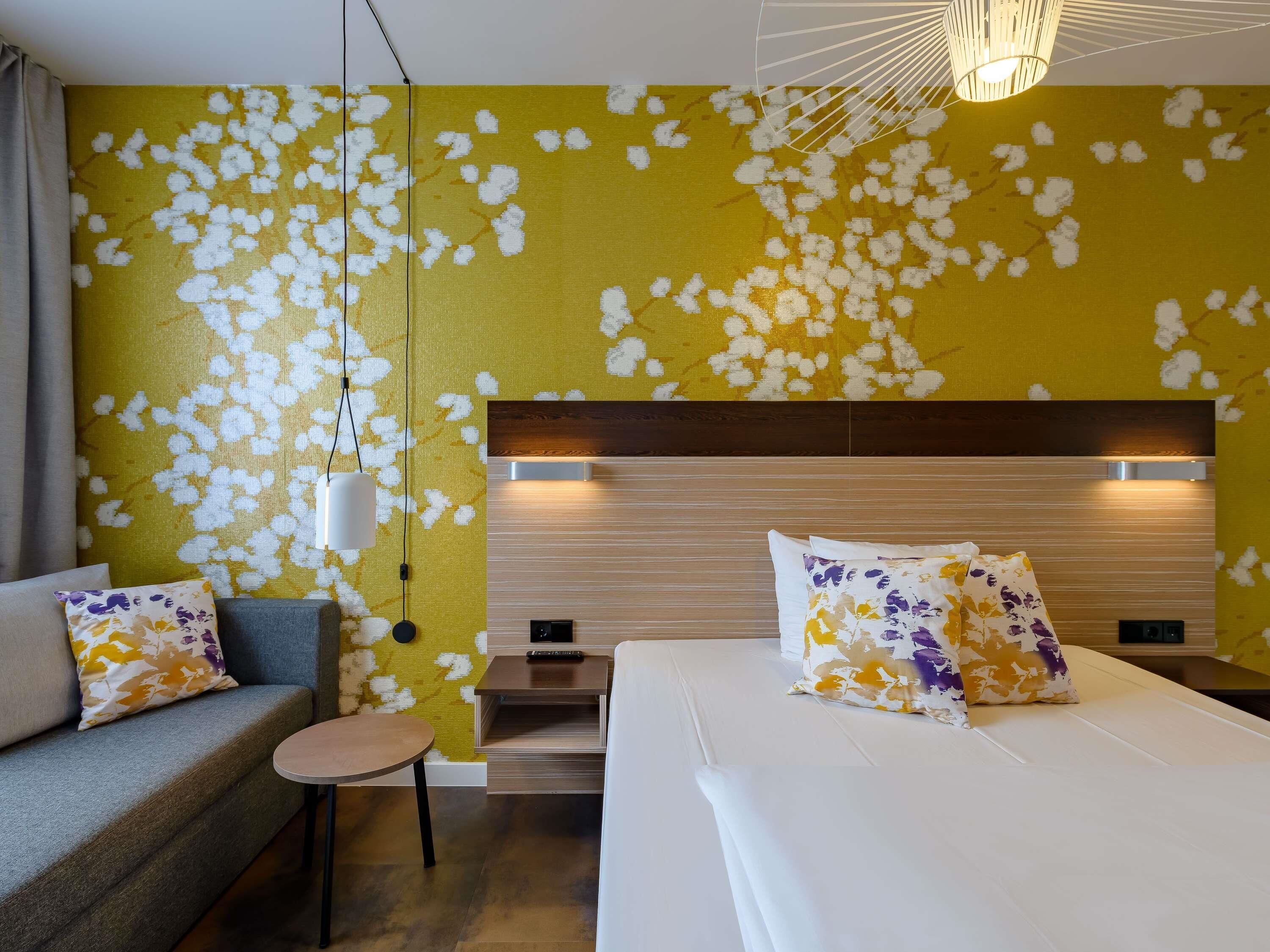 A hotel room with a bed, a sofa, a small table, and a wall with a yellow and white floral pattern.