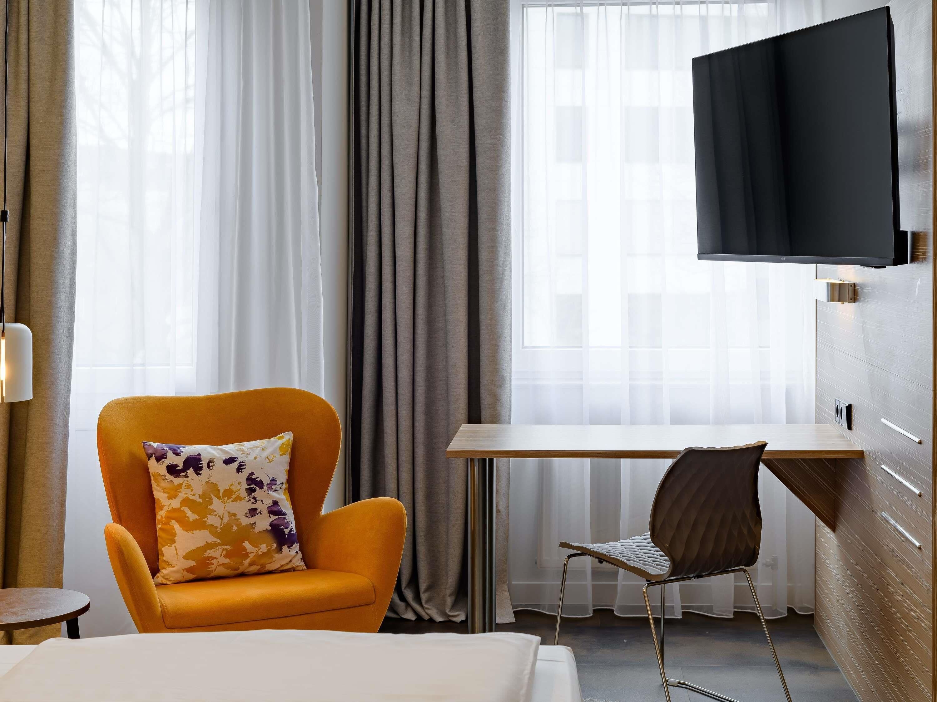 Een hotelkamer met een bed, een bureau, een oranje fauteuil, een bruine stoel en een flatscreen tv aan de muur.