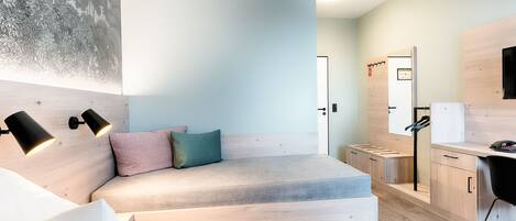 A modern bedroom with a wooden headboard, a bed with white and pink pillows, a desk with a chair, and a wall-mounted mirror.