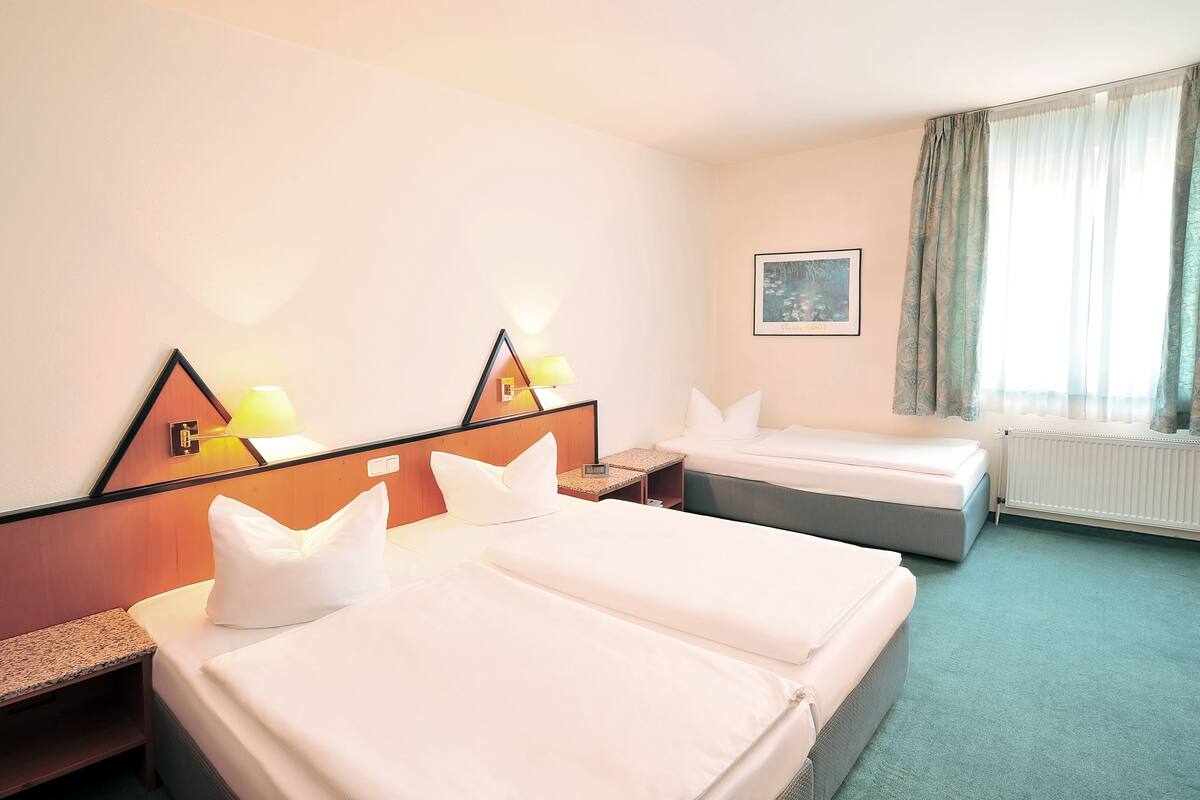 A hotel room with two beds, a nightstand, a lamp, a framed picture on the wall, and a window with curtains.