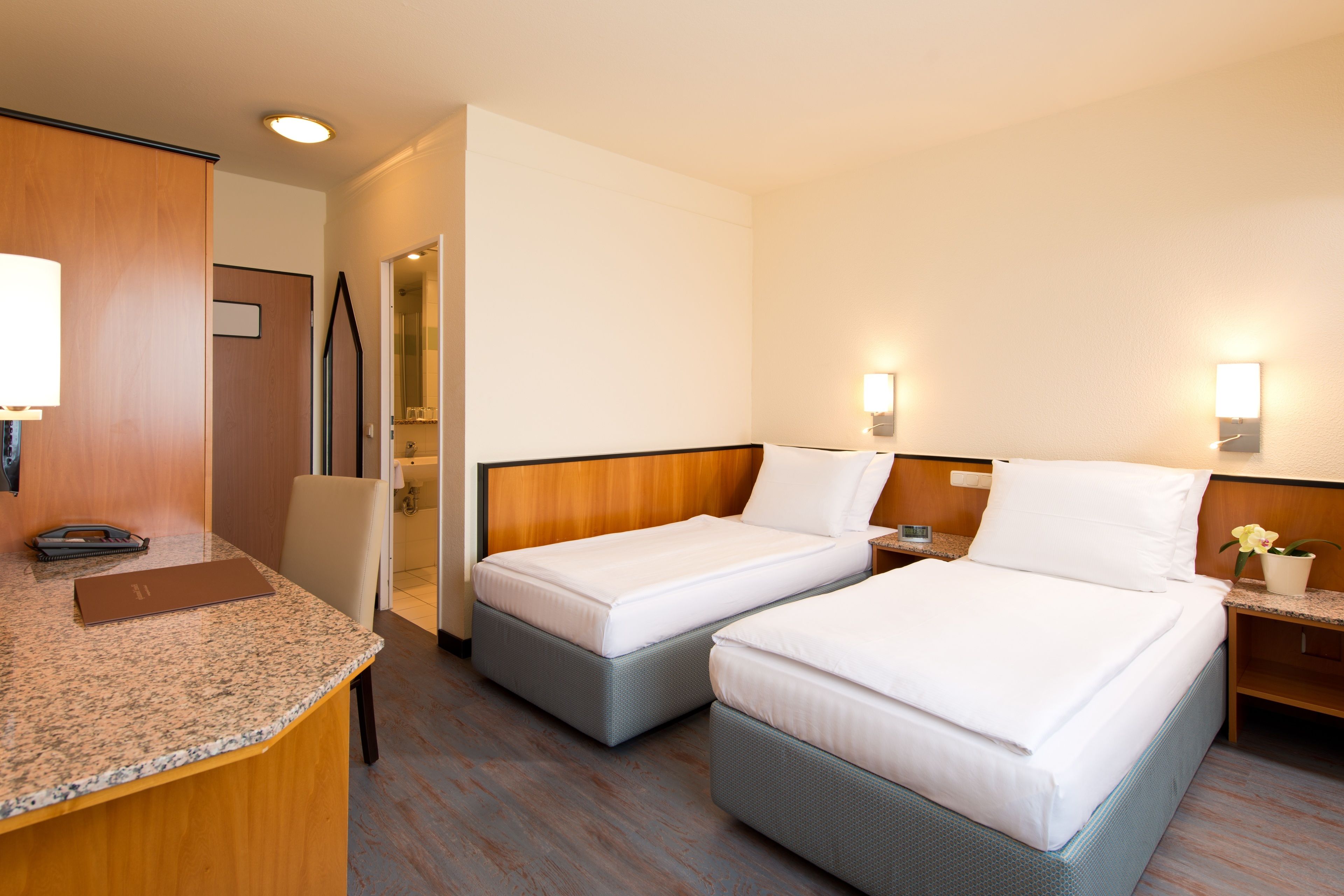 Ein Hotelzimmer mit zwei Einzelbetten, einem Schreibtisch mit Telefon und einem kleinen Tisch mit Blumenvase.
