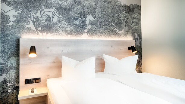 Una cama bien hecha con sábanas blancas, cabecera con acabado de madera y mesita de noche fijada a la pared con un interruptor y un objeto pequeño encima.
