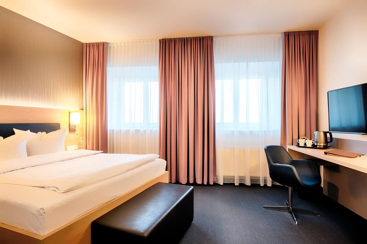 A hotel room with a bed, a desk, a chair, a TV, and a window with curtains.