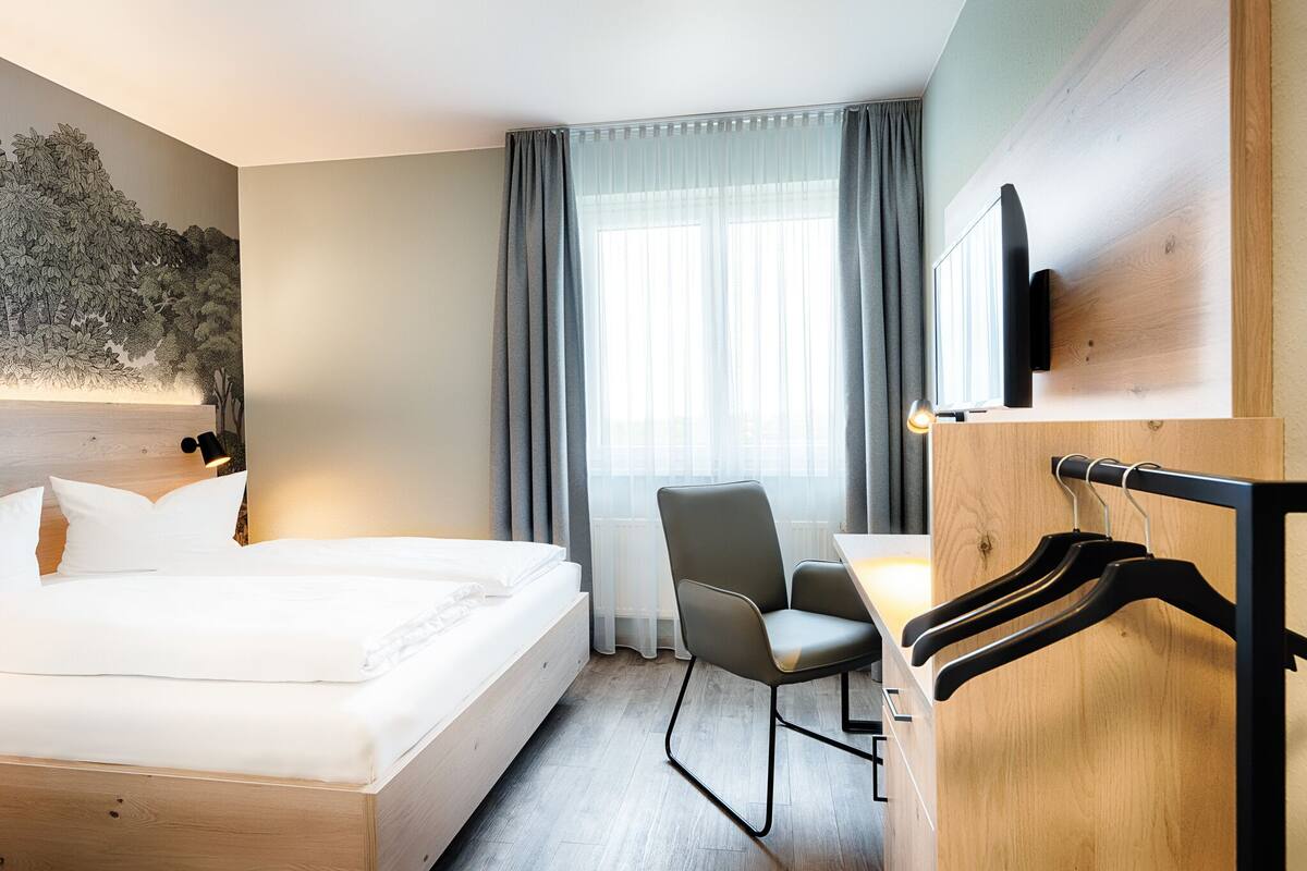 A hotel room with a bed, a desk, a chair, a TV mounted on the wall, and a coat rack.