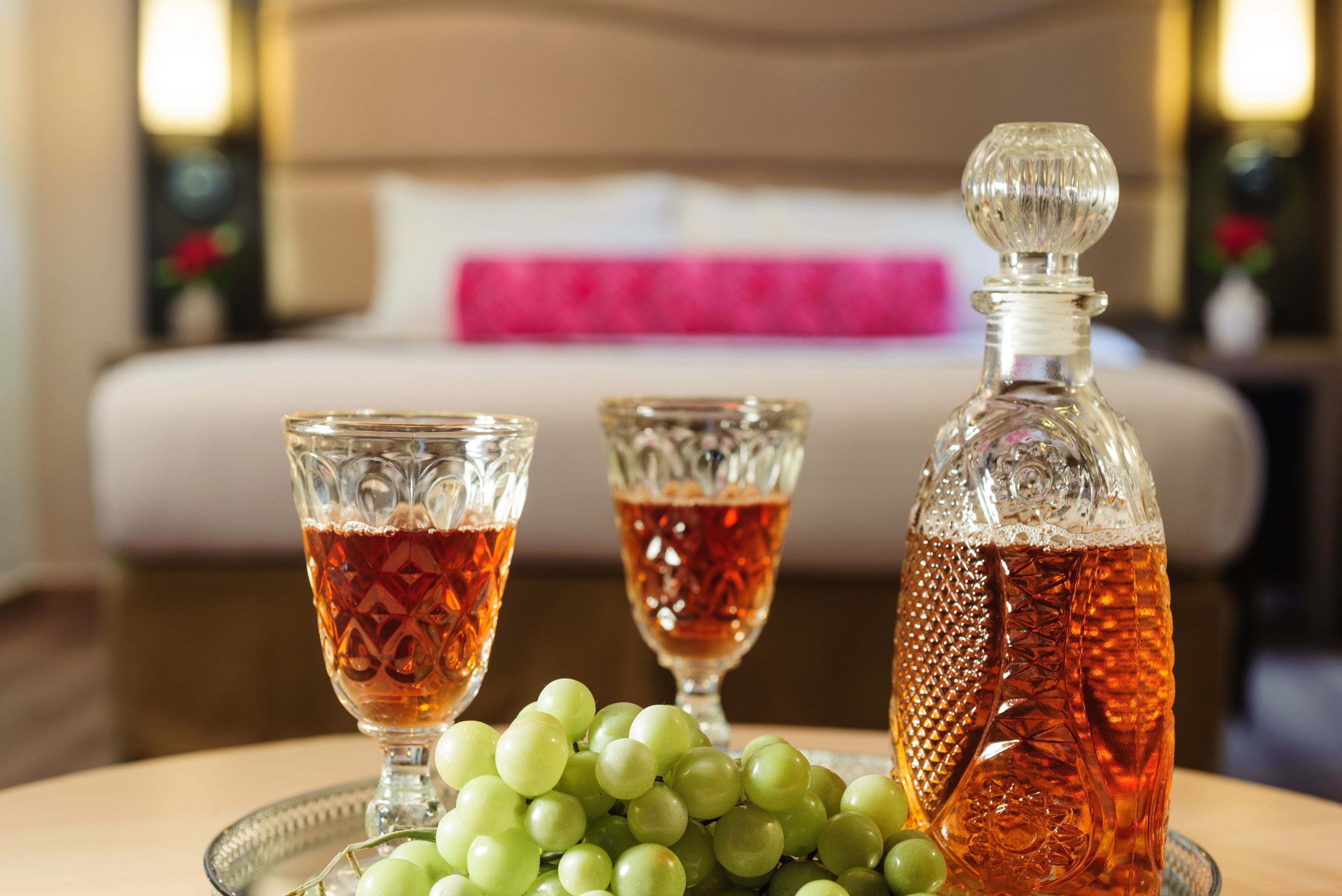 Una habitación con una cama, una mesita de noche con una botella y vasos de líquido ámbar, y una bandeja con uvas verdes.