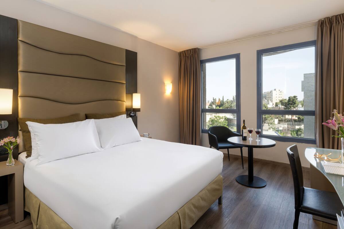 A hotel room with a large bed, a dining table with chairs, a view of the city through large windows, and a modern headboard.