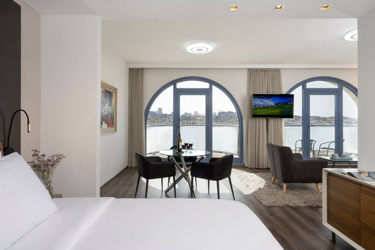 A hotel room with a bed, a dining table with chairs, a TV, and large arched windows with a city view.