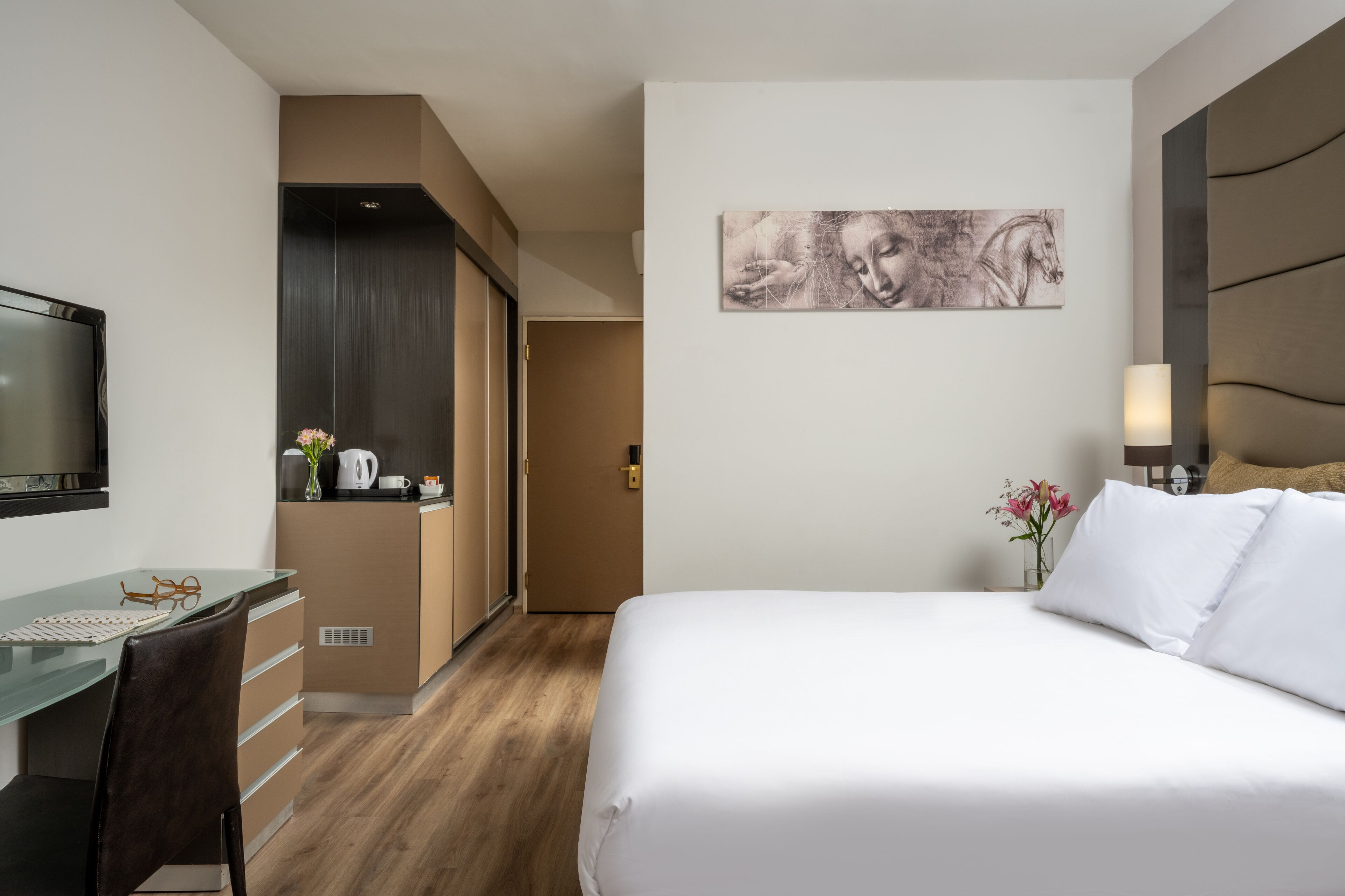 Een moderne hotelkamer met een bed, bureau en stoel, een flatscreen televisie, een schilderij aan de muur en een vaas met bloemen.