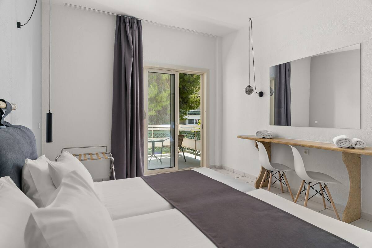 A modern hotel room with a bed, a desk with two chairs, a large mirror, and a sliding glass door leading to an outdoor area.