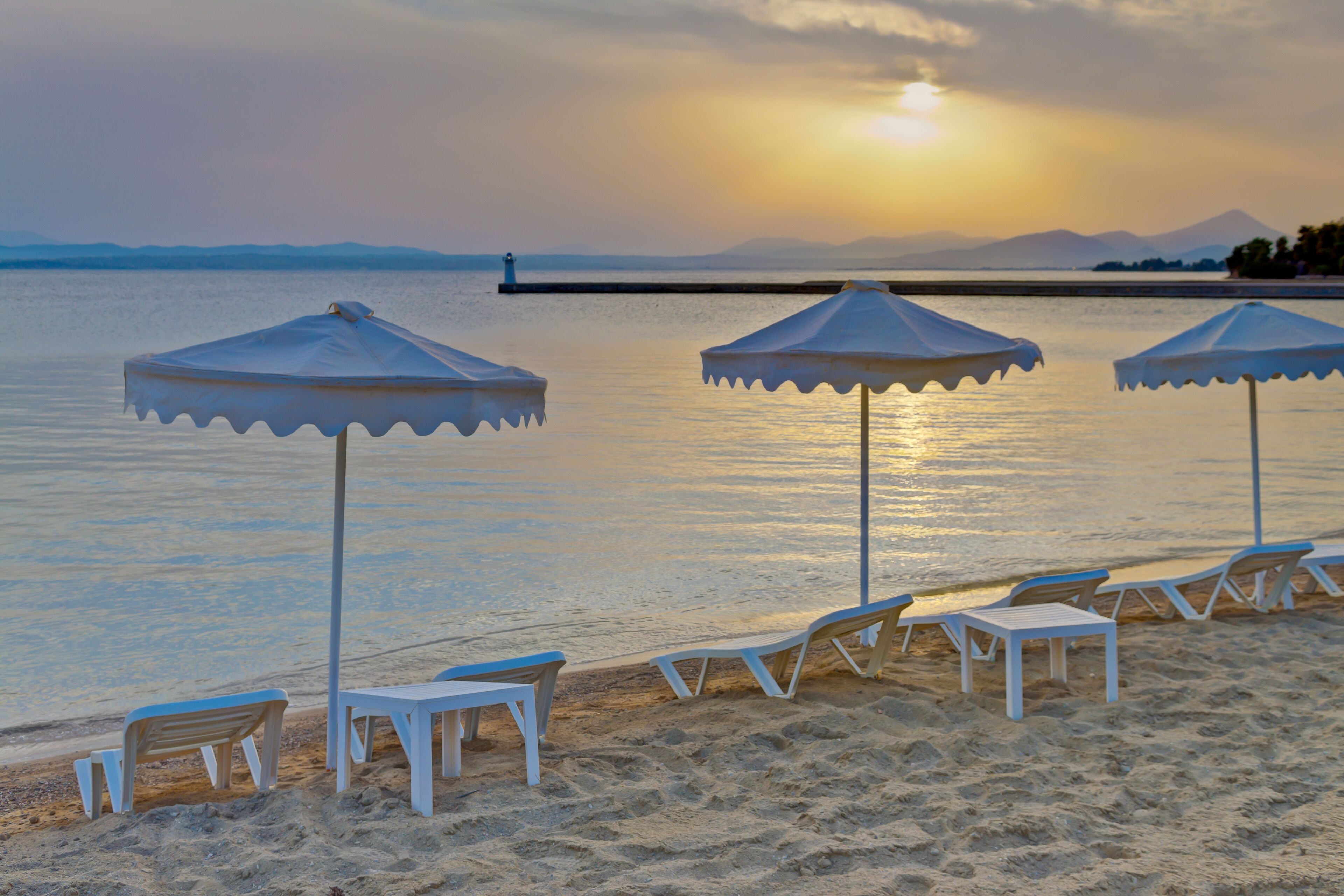 A beach with white umbrellas and lounge chairs, a calm sea, and a sunset in the background.