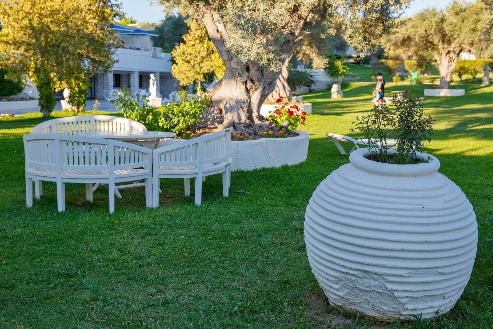 Un jardín con un banco blanco y una jardinera, un árbol y un edificio al fondo.